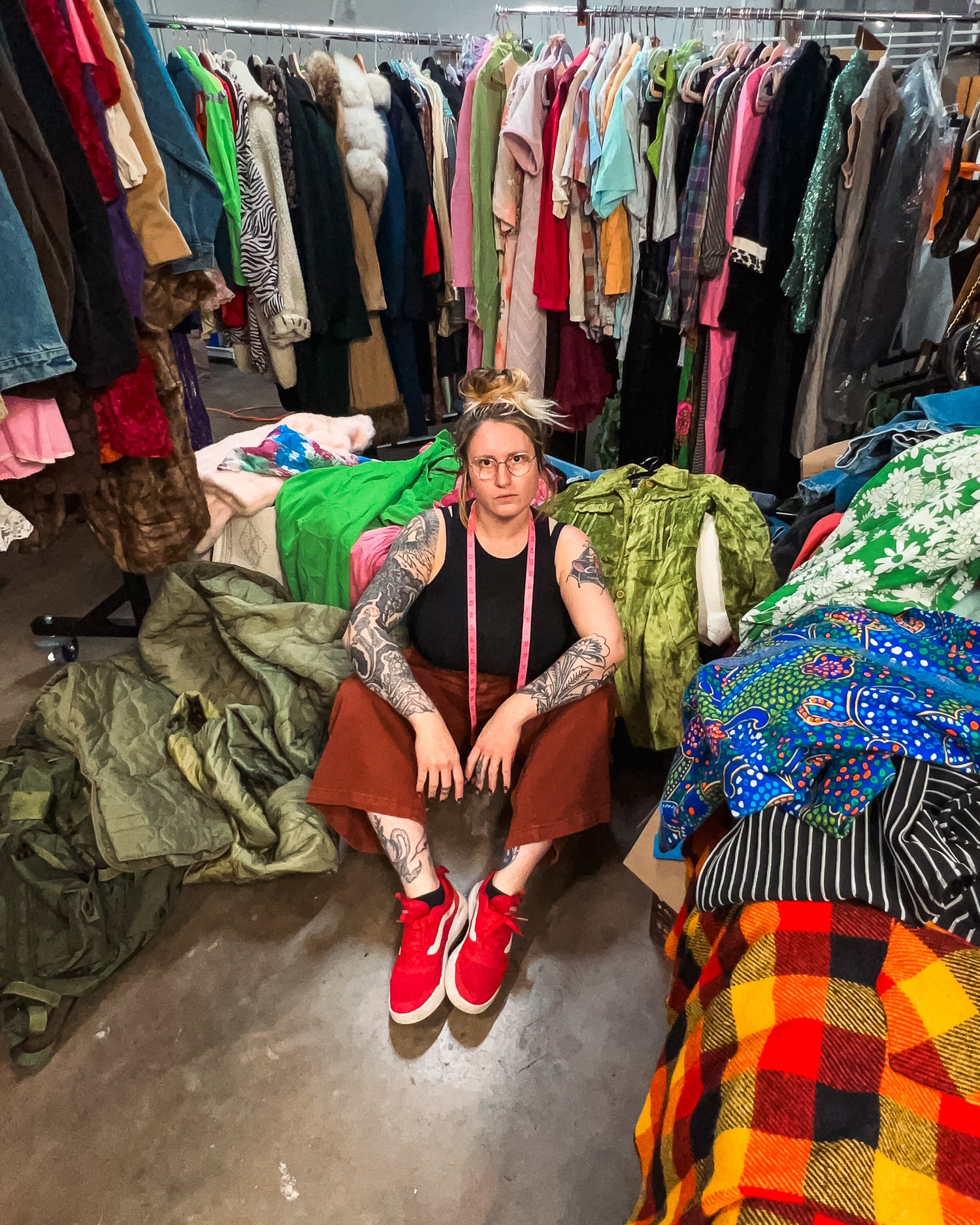Woman sitting on floor of warehouse surrounded by racks and piles of colorful vintage clothing. Taken from above looking down.